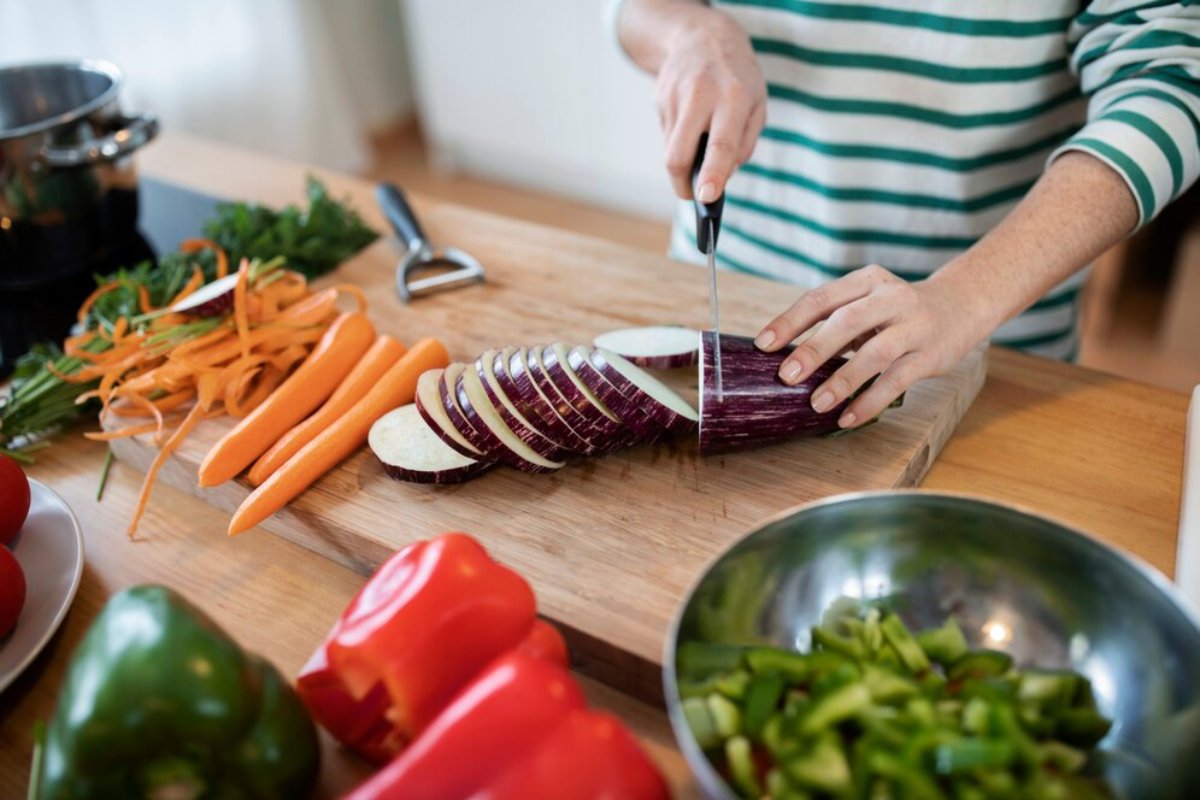 A person slices an eggplant on a wooden cutting board, surrounded by carrots, peppers, and herbs in a bright kitchen.