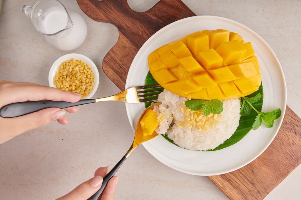 A close-up of a plate with sticky rice topped with mango, garnished with mint and cooked mung beans, with a hand holding utensils.