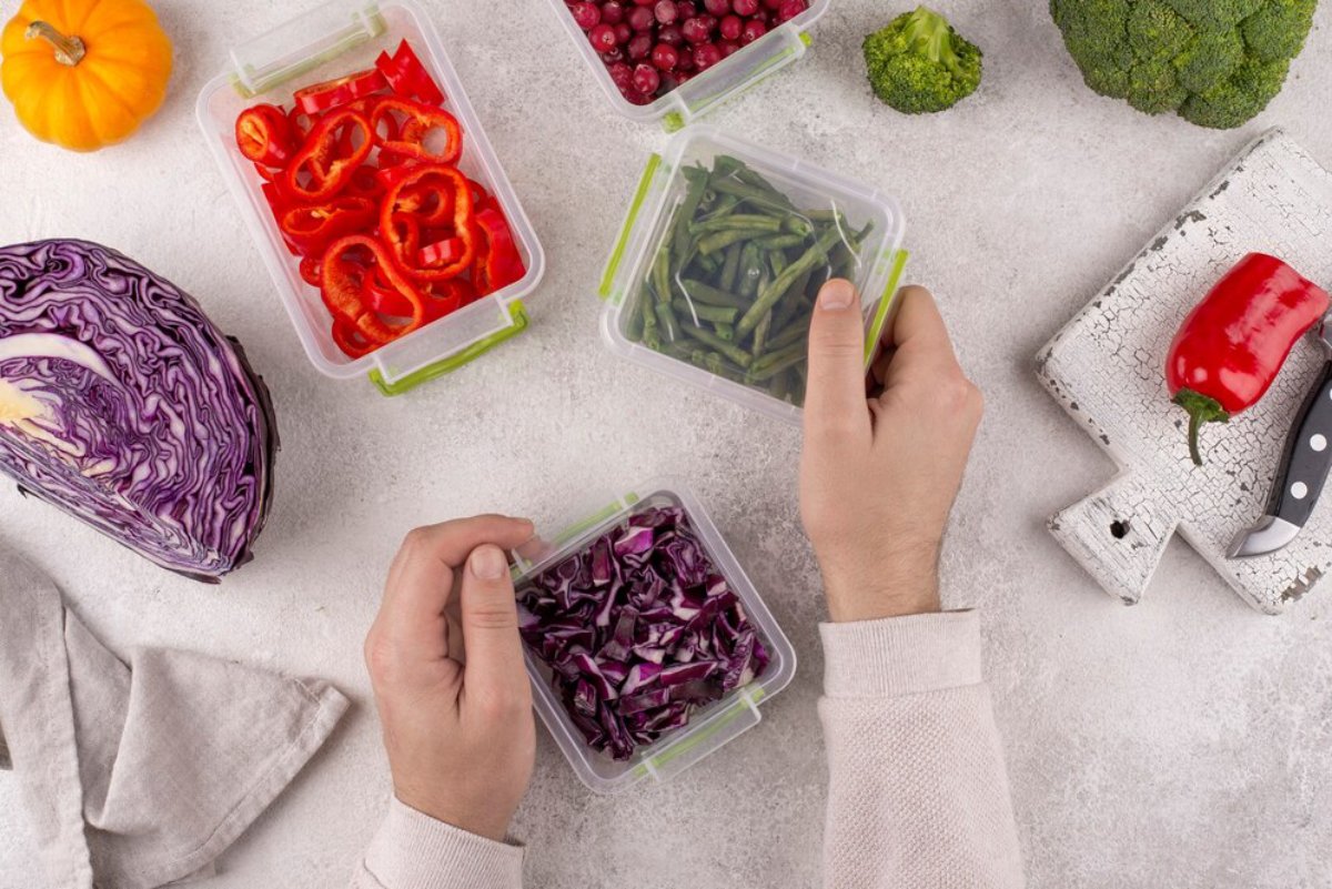 Two hands sorting fresh vegetables in clear containers on a kitchen surface with a pumpkin, red pepper, and knife nearby.