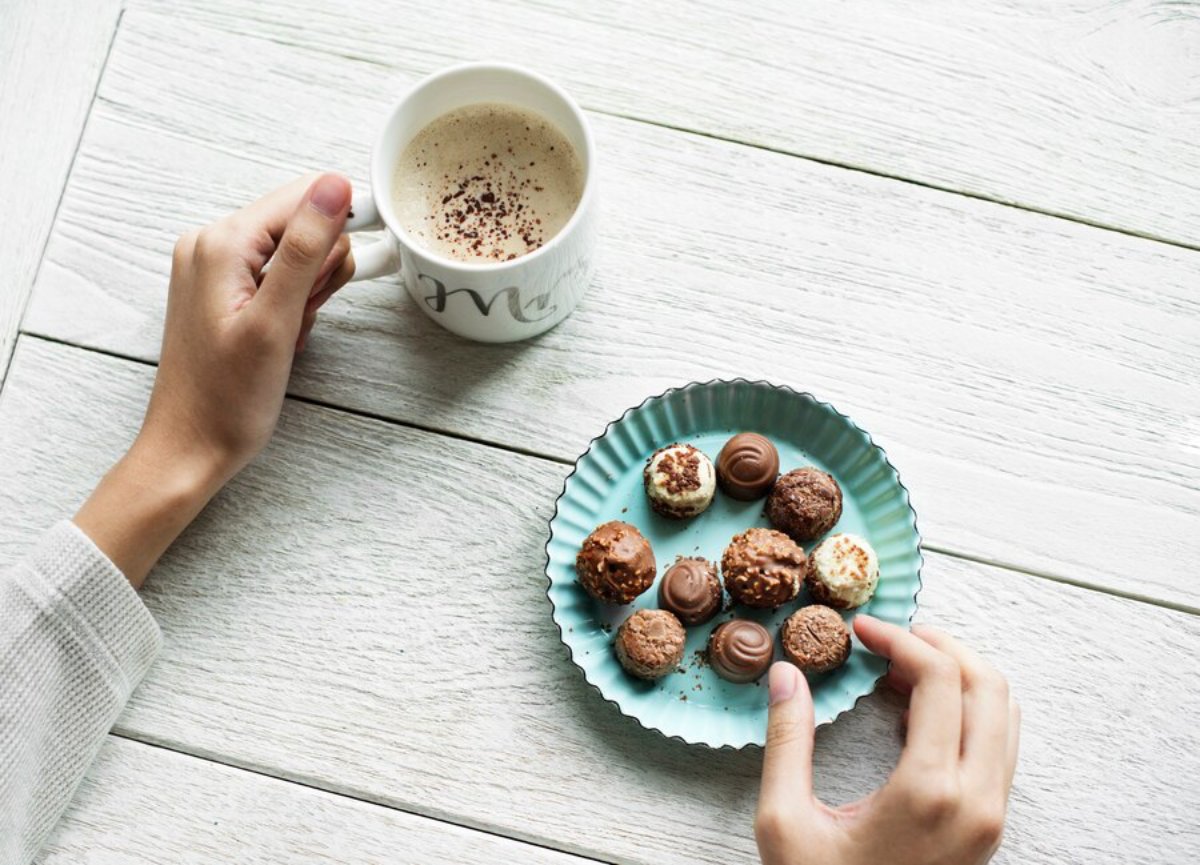 A hand holding a white mug of coffee next to a teal dish filled with assorted chocolate truffles on a light wooden table.
