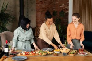 Three women prepare a vibrant spread of appetizers, including salads, olives, and tomatoes, in a stylish indoor setting.