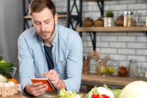 A person in a blue shirt writes in a notebook, surrounded by fresh vegetables and kitchen items on a wooden table.